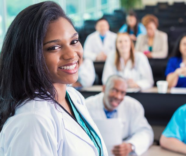 African American doctor or medical professional attending healthcare conference Young adult African American female doctor is standing in hospital lecture hall meeting room. She is attending medical conference or healthcare class with diverse doctors, nurses, and medical students. Adults are wearing scrubs and lab coats.